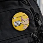 Racism Is Not Patriotism badge- Cats Against Fascism- Racism is not Patriotism Button Pin Badge, UK Political Merch, Yellow Badge with angry cat saying "Racism is not Patriotism" with Cats Against Fascism Text and Logo above Mock up on Black back pack