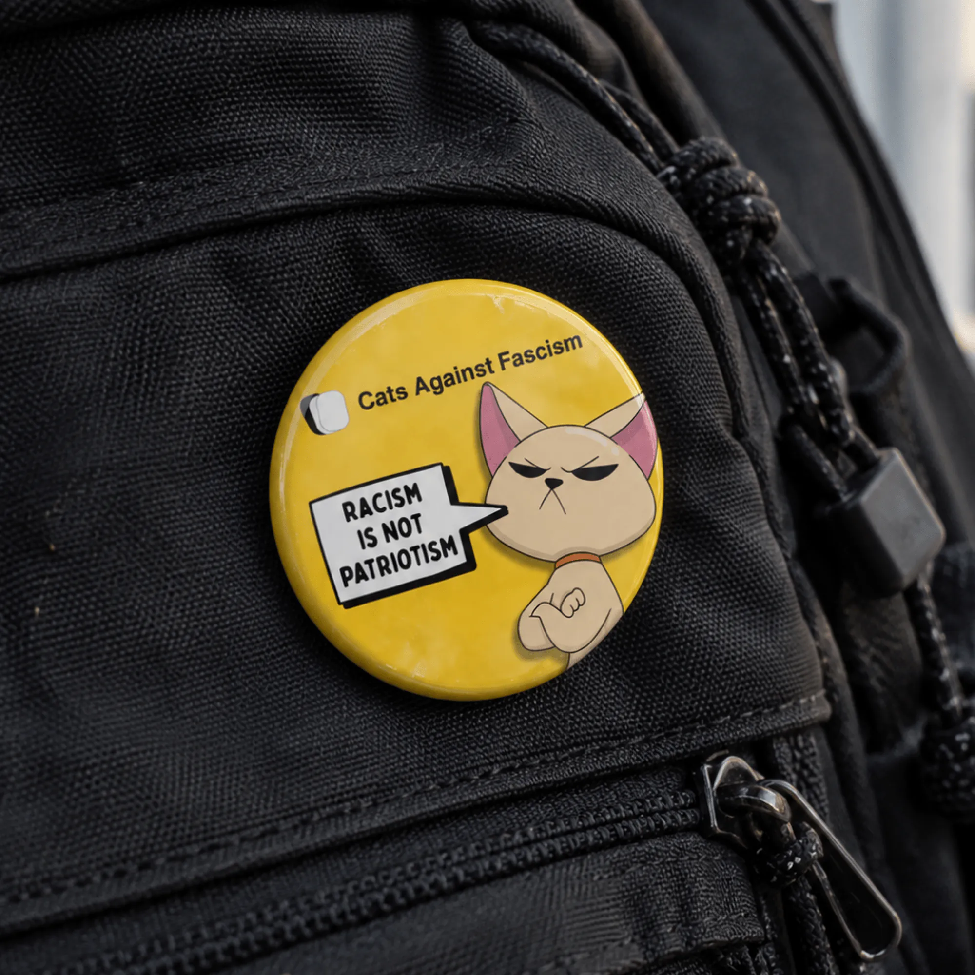 Racism Is Not Patriotism badge- Cats Against Fascism- Racism is not Patriotism Button Pin Badge, UK Political Merch, Yellow Badge with angry cat saying "Racism is not Patriotism" with Cats Against Fascism Text and Logo above Mock up on Black back pack