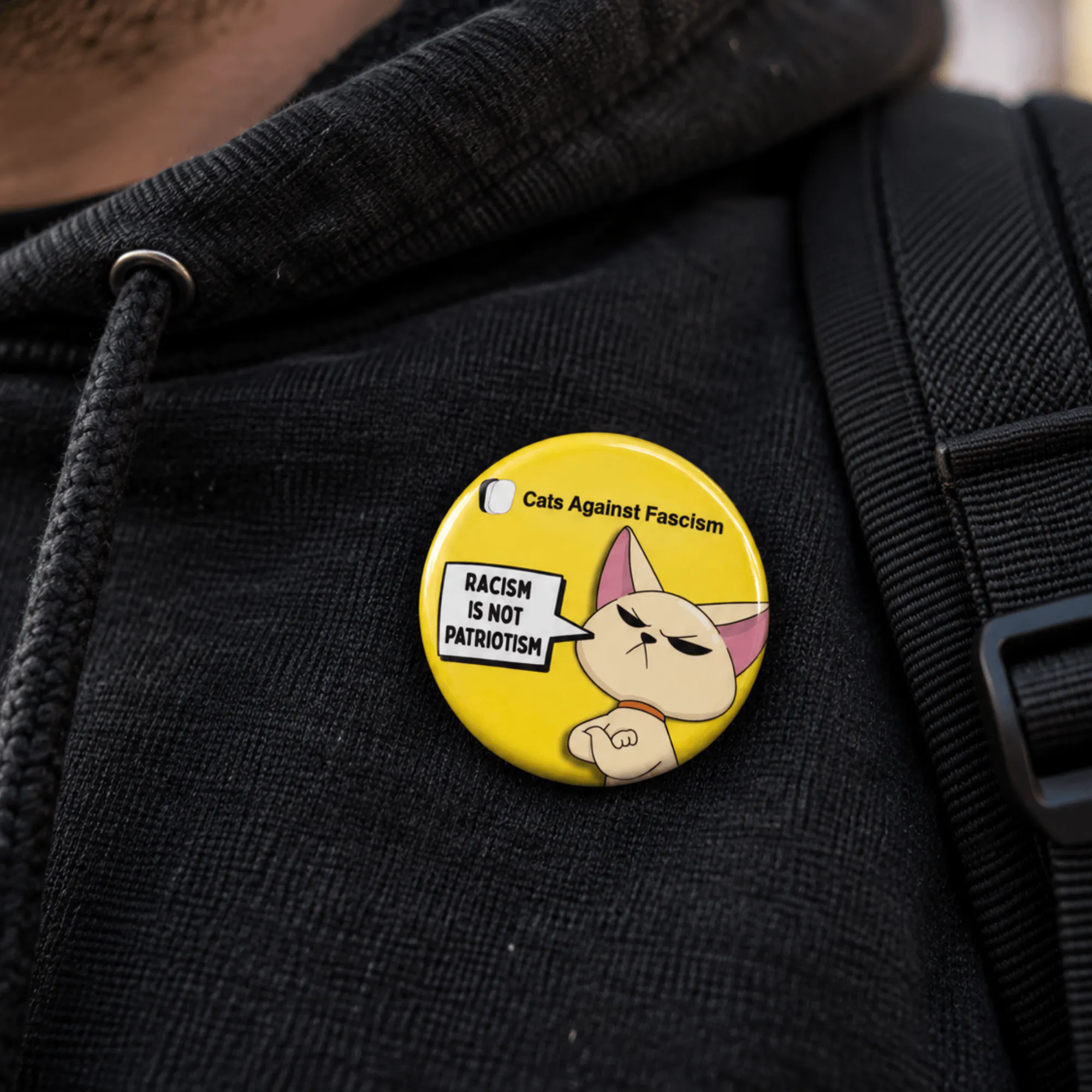 Racism Is Not Patriotism badge- Cats Against Fascism- Racism is not Patriotism Button Pin Badge, UK Political Merch, Yellow Badge with angry cat saying "Racism is not Patriotism" with Cats Against Fascism Text and Logo above Mock up on Black Hoody