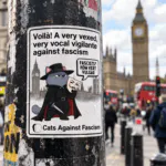 Guy Fawkes Cat Vinyl Sticker – Cats Against Fascism Vigilante cat sticker – grey cat in black hat and cloak holding a Guy Fawkes mask in front of Parliament