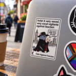Guy Fawkes Cat Vinyl Sticker – Cats Against Fascism Vigilante cat sticker – grey cat in black hat and cloak holding a Guy Fawkes mask in front of Parliament