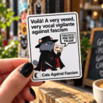 Guy Fawkes Cat Vinyl Sticker – Cats Against Fascism Vigilante cat sticker – grey cat in black hat and cloak holding a Guy Fawkes mask in front of Parliament